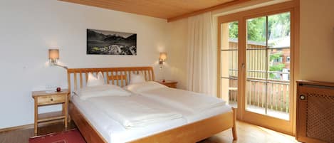 A bedroom with a wooden bed, two bedside tables, a nightstand, a red rug, a large window with a view, and a wall-mounted picture.