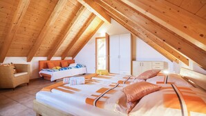 Un dormitorio cálido en el ático, con techo inclinado, una cama grande con ropa de cama naranja, una silla de mimbre y un armario.