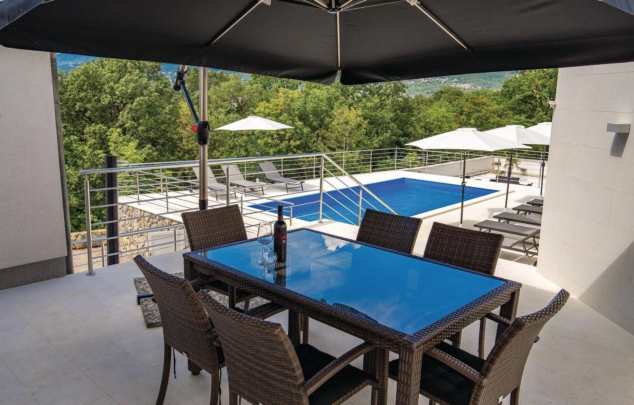 Een zwembadterras met een glazen tafel en rieten stoelen, omringd door parasols en uitzicht op een zwembad en begroeiing.