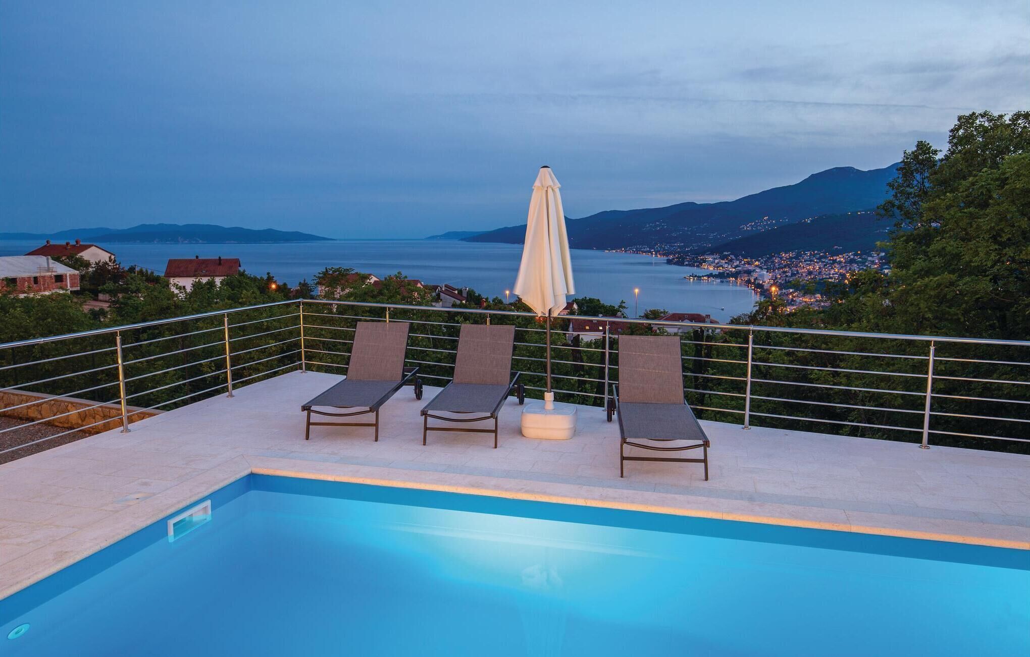 A poolside area with two lounge chairs and a table, overlooking a scenic view of a lake and distant mountains.
