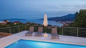 Ein Poolbereich mit zwei Liegestühlen und einem Tisch, mit Blick auf eine malerische Seenlandschaft und ferne Berge.