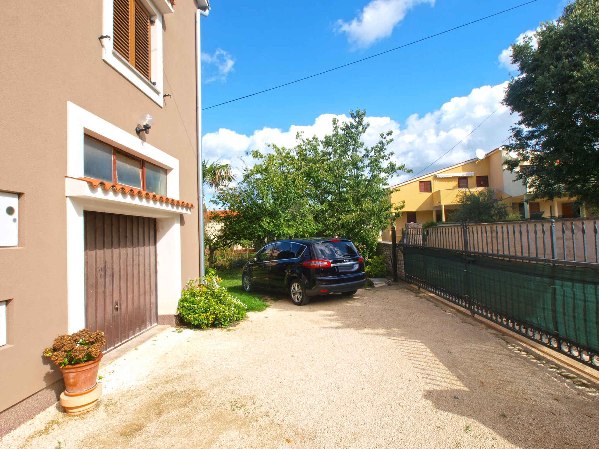 Rua residencial com um carro estacionado na entrada, uma casa com fachada marrom e cerca verde.