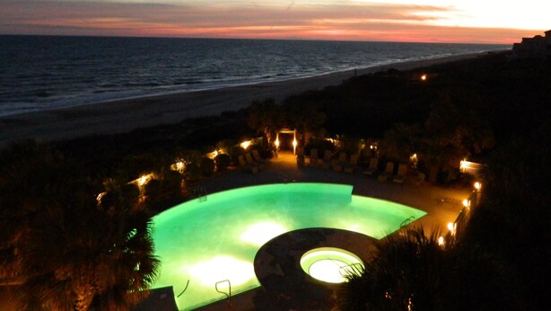 A swimming pool with a circular hot tub, surrounded by lounge chairs, set against a sunset beach view. - Beach Front Condo With Wonderful Ocean View  (Indian Beach)