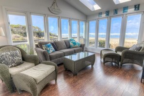 A sunlit room with a large window overlooking a beach, furnished with wicker furniture and a glass coffee table.