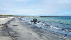 Une plage de sable avec un grand rocher dans l’eau, un ciel dégagé et une côte lointaine.