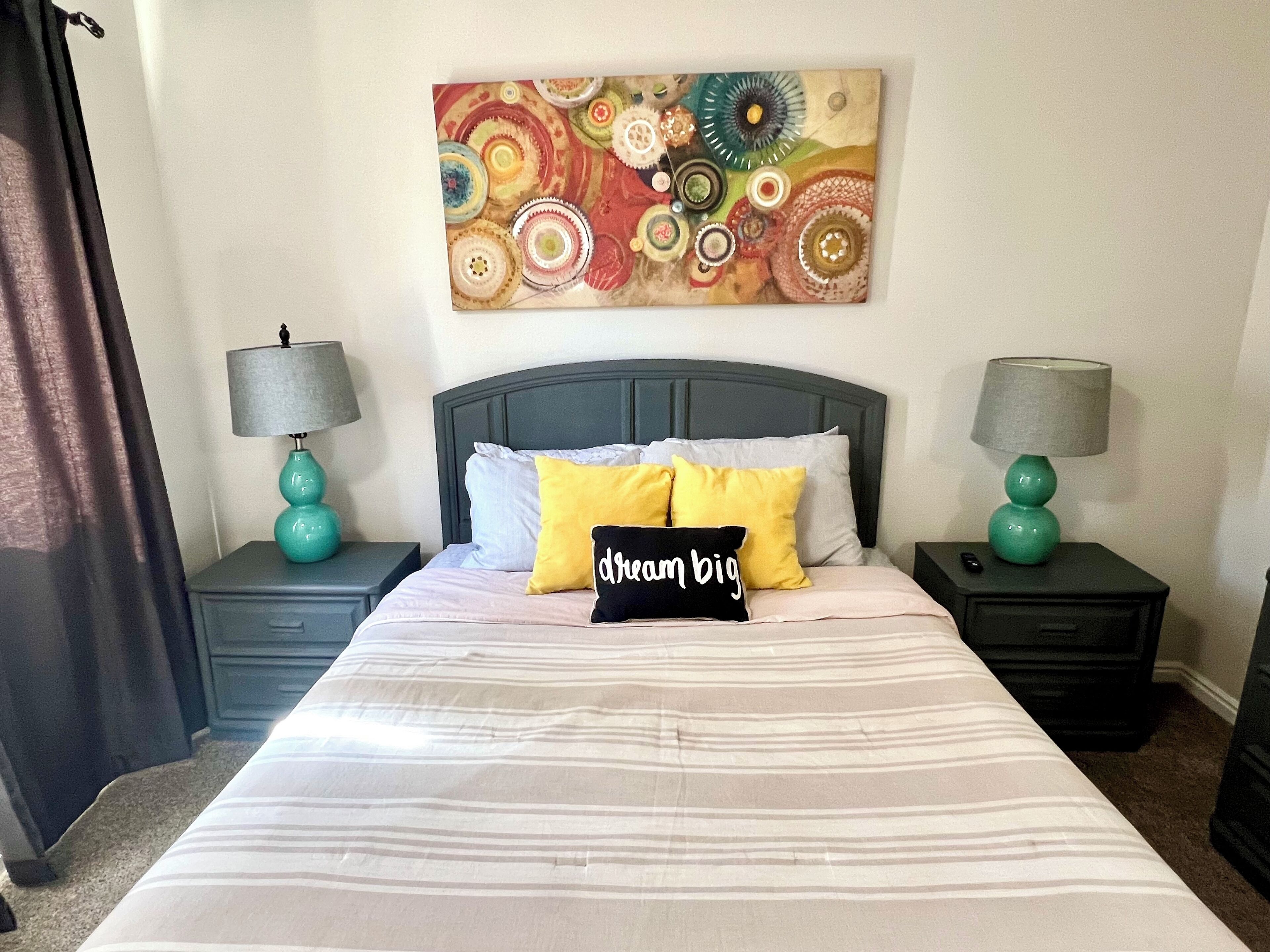 A bed with striped bedding, two bedside tables with lamps, and a colorful abstract painting on the wall.