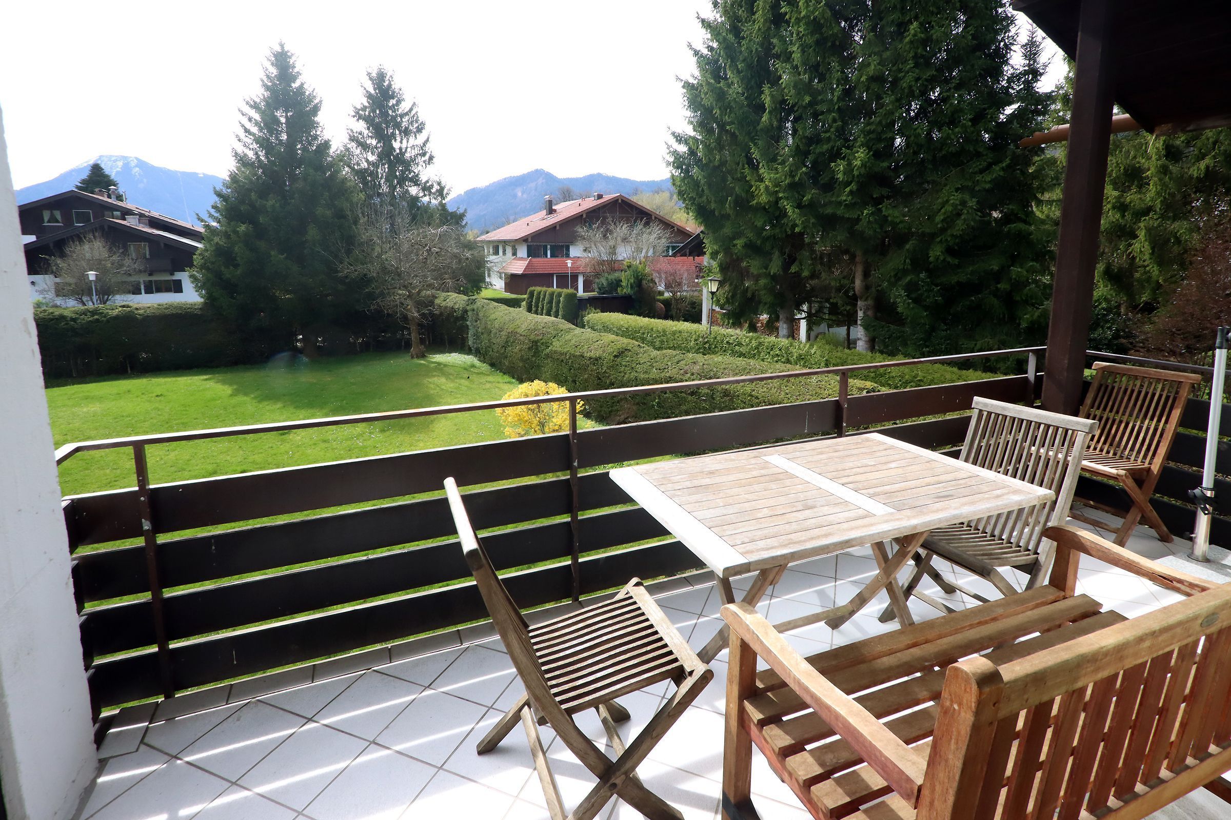 Ein Balkon mit Tisch und Stühlen, Blick auf einen grünen Garten und Häuser im Hintergrund.
