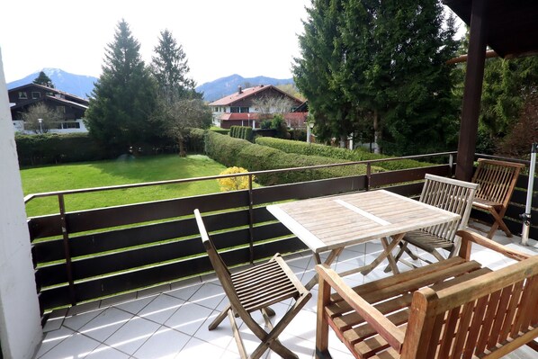 Een balkon met een tafel en stoelen, uitzicht op een groene tuin en huizen op de achtergrond.