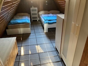 Ein schmales Zimmer mit zwei Betten, einem kleinen weißen Schrank und einem gefliesten Boden.