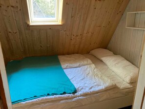 Pequeña habitación individual con paredes de madera, una ventana y una cama sencilla con una manta verde azulada.