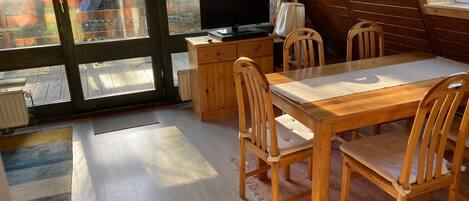 Ein gemütliches Zimmer mit einem Holzesstisch, Stühlen, einem Fernseher und einem großen Fenster.