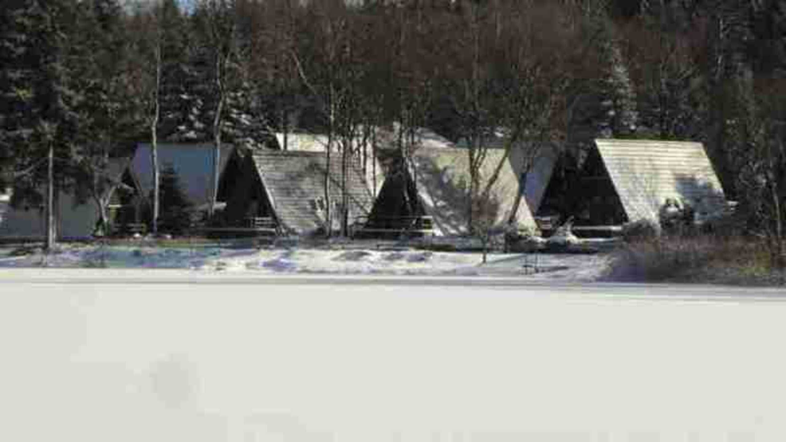A row of simple, single-story cabins with sloped roofs, surrounded by trees and a snowy landscape.