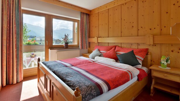 A bedroom with a wooden headboard, a large window with curtains, a bed with multiple pillows, and a nightstand with a vase.