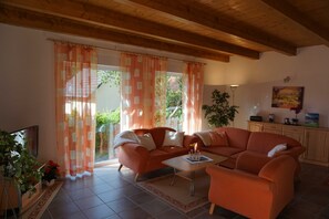 A living room with a sofa, a coffee table, and a TV. There are curtains, a plant, and a painting on the wall.