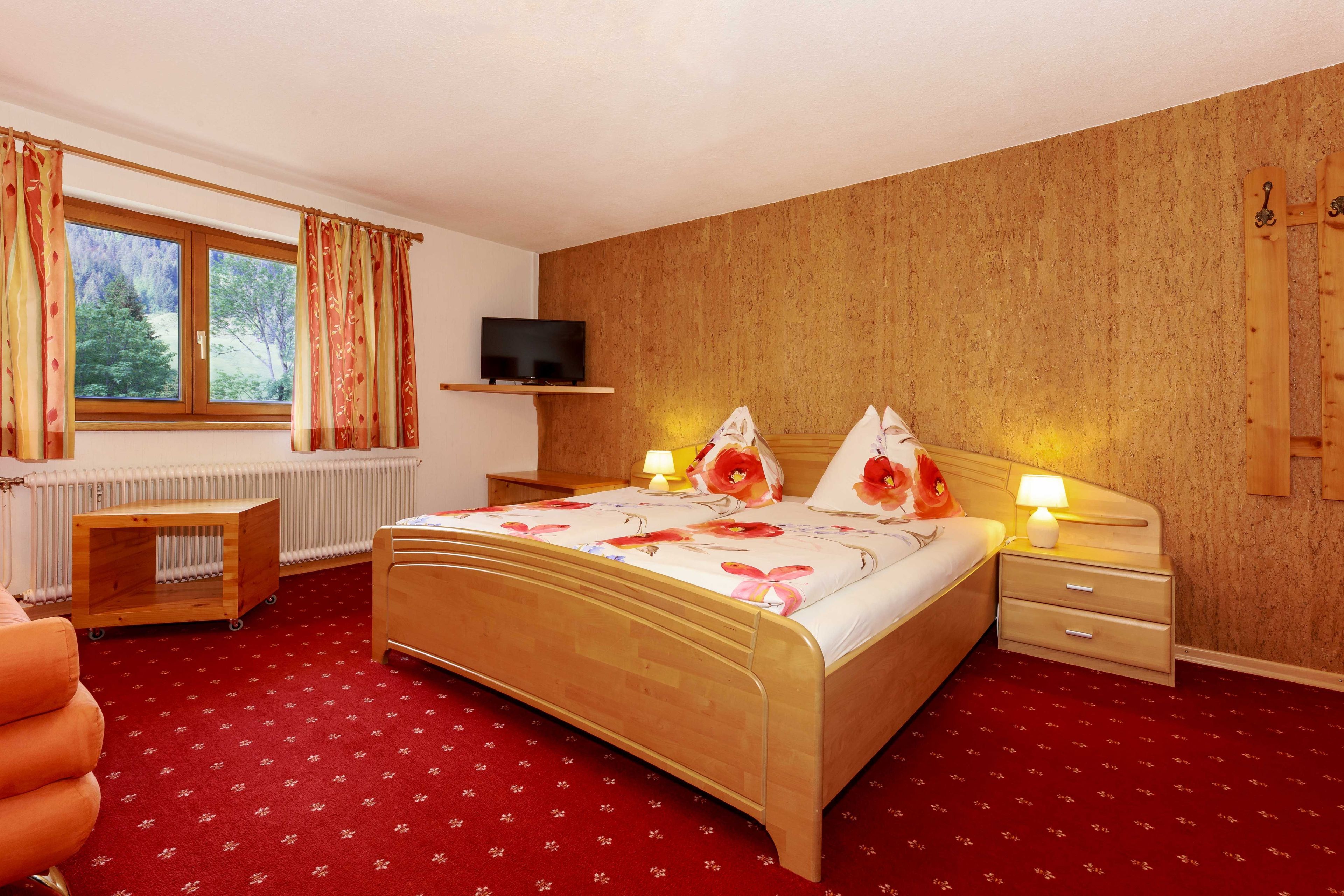 A bedroom with a wooden bed, a bedside table, a nightstand, a TV on a shelf, a radiator, and a window with curtains.
