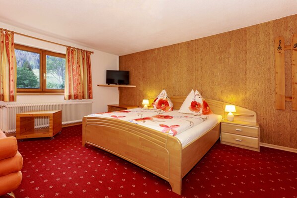 A bedroom with a wooden bed, a bedside table, a nightstand, a TV on a shelf, a radiator, and a window with curtains.