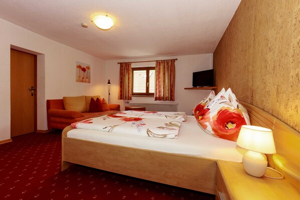 Quarto de hotel com cama, sofá, televisão, janela com cortinas e abajur.