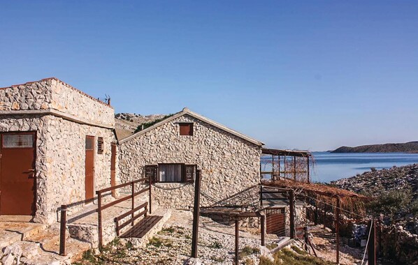 A stone building with a wooden door and a small window, situated near a rocky coastline with a clear blue sky.