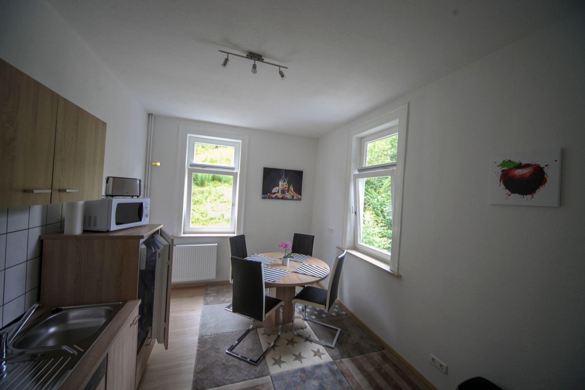A kitchen with a microwave, a sink, and a dining area with a table and chairs. There are two windows providing natural light.