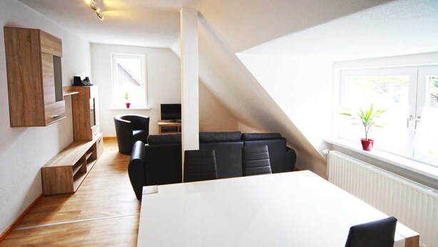 A modern interior with a large white table, black chairs, a wooden cabinet, and a view through a window. - Apartment Lerbachtal - Apartment Lerbachtal L3 2-6 persons (Osterode am Harz)