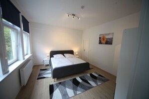 A bedroom with a bed, bedside tables, a window, a radiator, a ceiling light, and a painting.