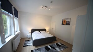 A bedroom with a bed, bedside tables, a window, a radiator, a ceiling light, and a painting. - Apartment Lerbachtal - Apartment Lerbachtal L3 2-6 persons (Osterode am Harz)
