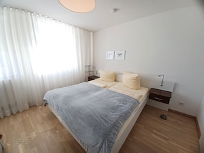 A bedroom with a bed, a bedside table, a nightstand, a window with curtains, and a wall-mounted light fixture. - Käptn Brass App 27 - Käpt`n Brass App.27 (Dierhagen)