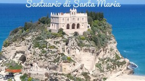 A historic building with a prominent bell tower, surrounded by a rocky landscape and palm trees, overlooking a coastal area.