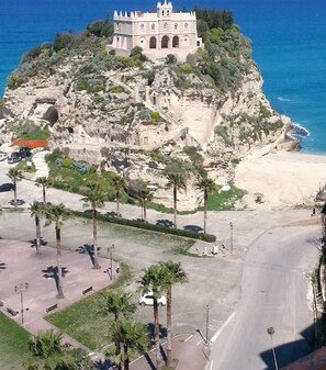 Un edificio histórico con una imponente torre campanario, rodeado de un paisaje rocoso y palmeras, con vistas a la costa.