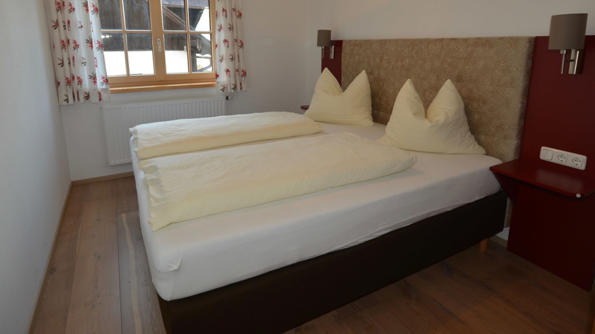 Una cama con dos almohadas, suelo de madera, ventana con cortinas florales y una lámpara fijada a la pared.