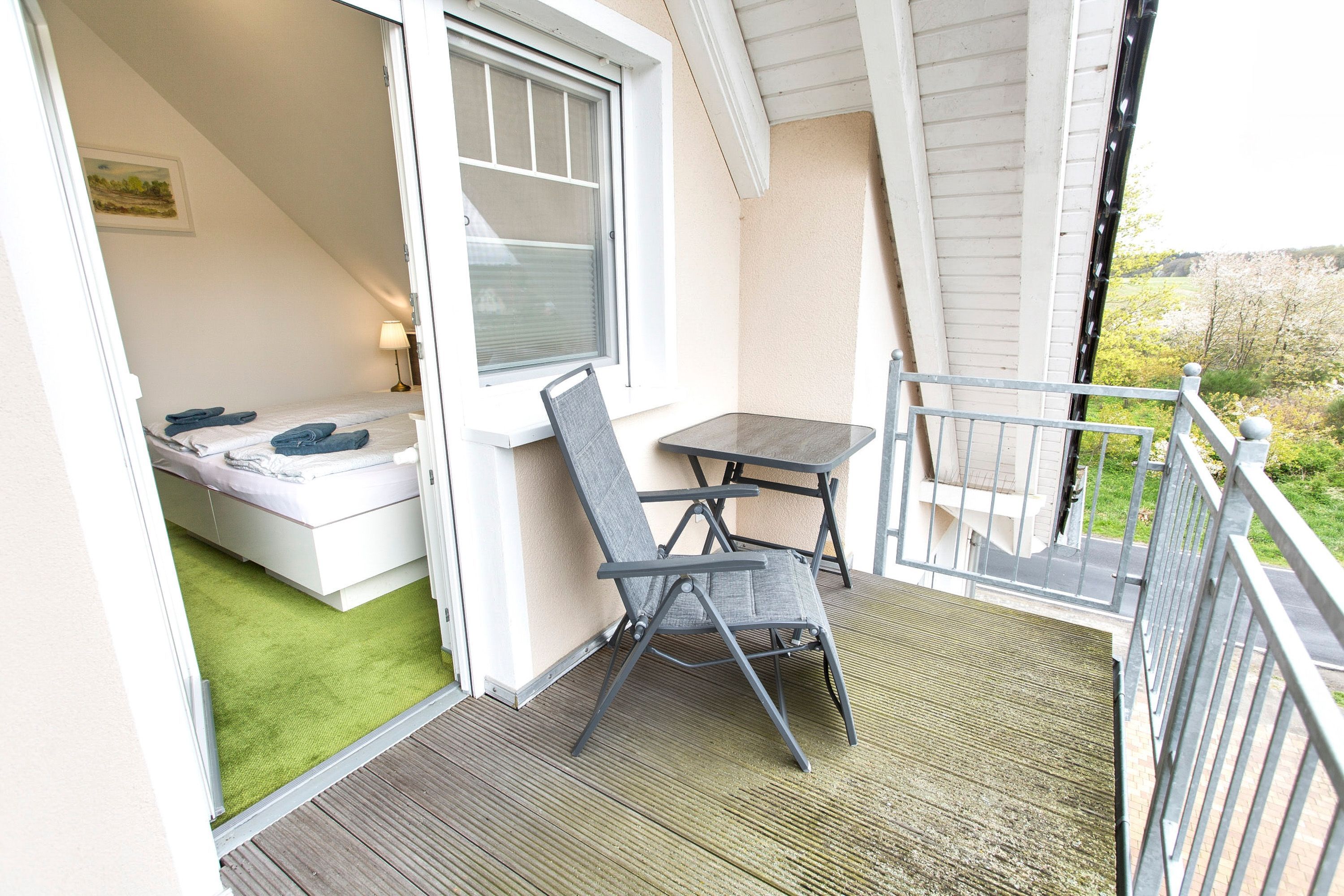 A balcony with a table and chair, a bed with a green mat, and a window with blinds.