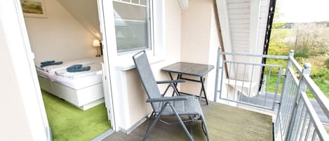 A balcony with a table and chair, a bed with a green mat, and a window with blinds.