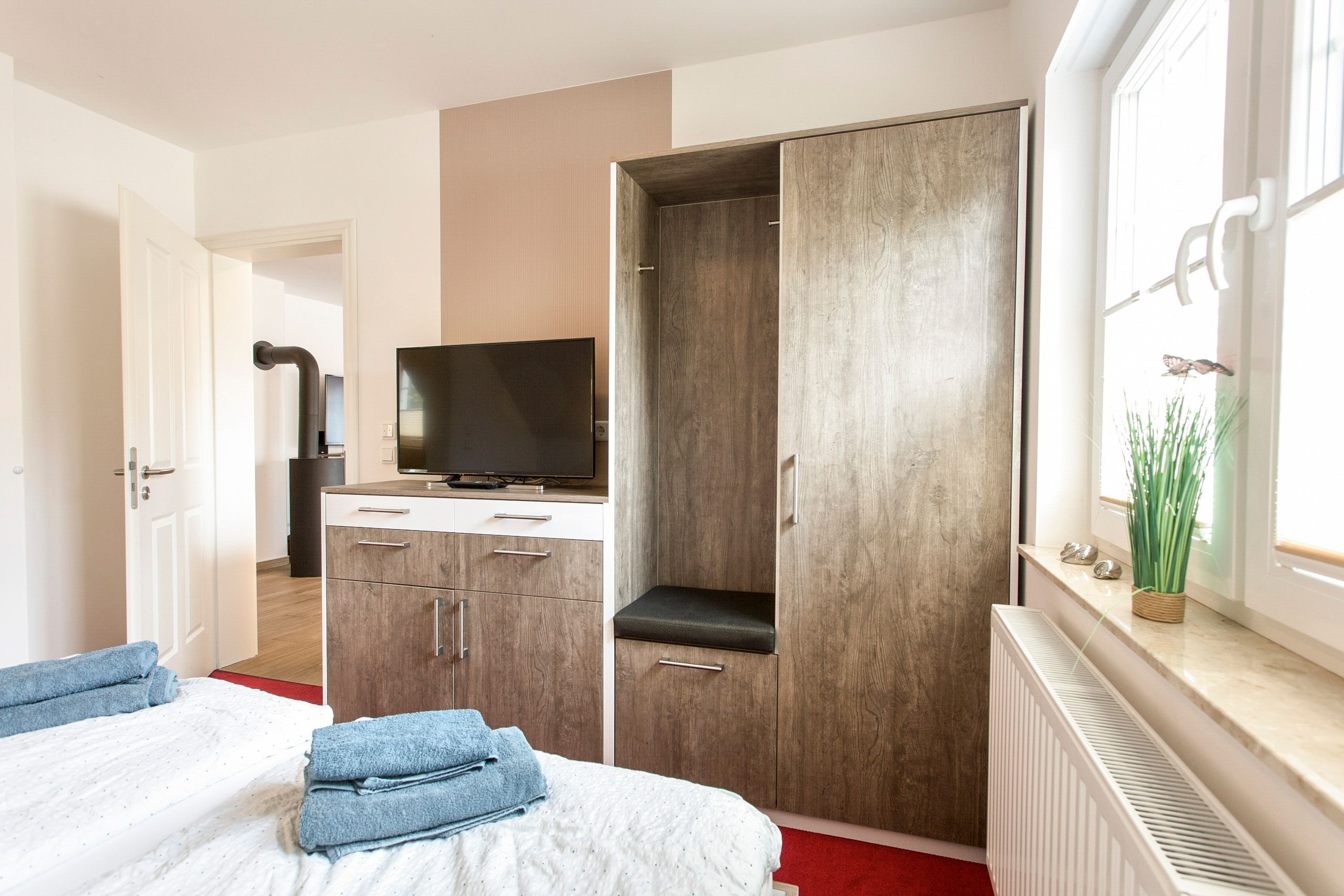 A bedroom with a bed, a wardrobe, a television, and a window with plants.