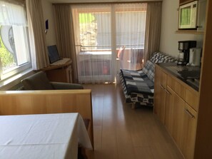 A compact room with a bed, a small table, a kitchenette, and a sliding glass door leading to an outdoor area.