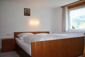 A bedroom with a wooden bed, a bedside table, a wall-mounted light, and a window with a view of the outdoors.