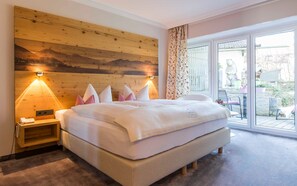 Ein Hotelzimmer mit Holz-Kopfteil, einem großen Bett, zwei Nachttischlampen, einem kleinen Nachttisch und Blick durch eine Glastür nach draußen.