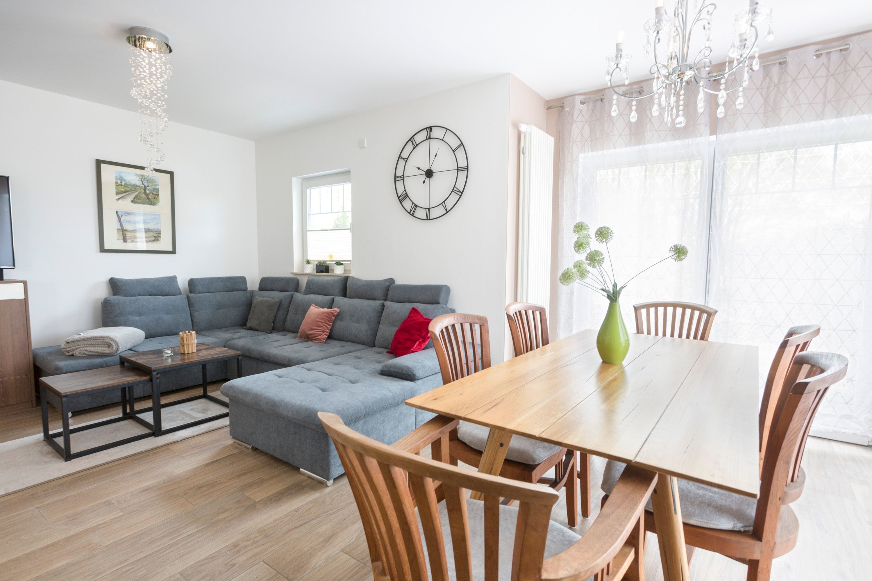 Ein modernes Wohnzimmer mit einem grauen Ecksofa, einem Holzesstisch mit Stühlen, einer Wanduhr und einer Pendelleuchte.