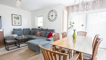 Ein modernes Wohnzimmer mit einem grauen Ecksofa, einem Holzesstisch mit Stühlen, einer Wanduhr und einer Pendelleuchte.