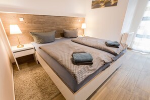 A modern bedroom with a wooden headboard, two bedside tables with lamps, a large bed with patterned bedding, and a view of the outdoors through a window.