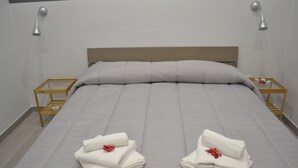 A neatly made bed with white linens and two folded towels with red flowers on the bed. There are two bedside tables with lamps and a wooden nightstand with a glass container. - Bassetto 013 (Naples)