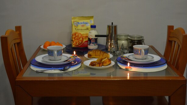 A dining table set with plates, cups, and food items, including a croissant and a bag of cereal. - Bassetto 013 (Naples)