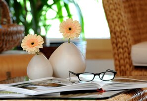 Um livro com óculos por cima, dois vasos com flores e uma cadeira de vime ao fundo.