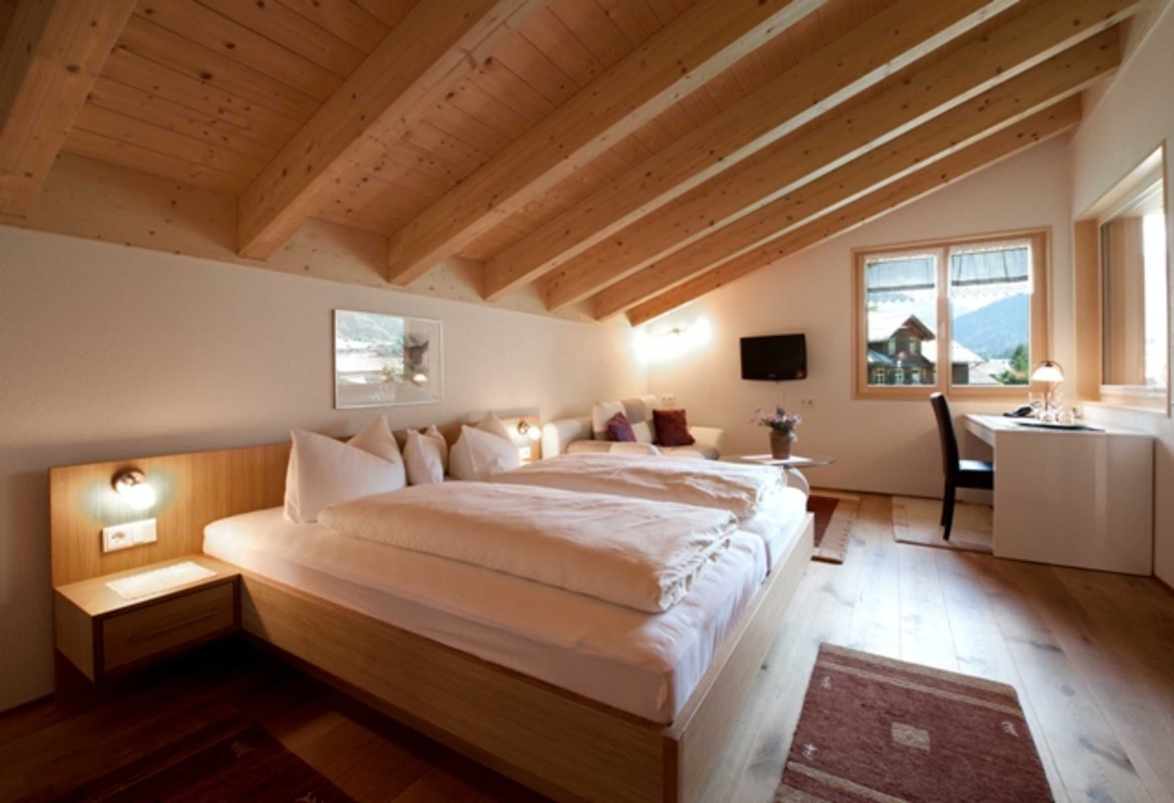 Une chambre avec un plafond en bois, un lit avec une literie blanche, une table de chevet avec une lampe, un bureau avec une chaise et une fenêtre donnant sur une montagne.