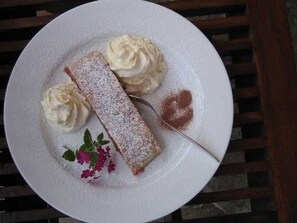 Un plato de postre con un pastel, crema batida y una pizca de azúcar glas, acompañado de una ramita de flores.