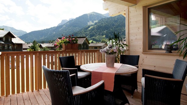 A wooden deck with a table and chairs, a view of mountains, and a window with a view of a house.