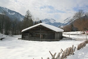 A wooden cabin with a snow-covered roof, surrounded by a wooden fence, set against a backdrop of snow-covered mountains and trees. - Category B - Room Künzelblick - WohlfühlHaus Evelin (Au)