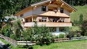 A two-story chalet with a balcony, surrounded by lush greenery and a wooden fence.