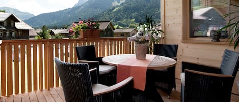 A wooden deck with a table and chairs, a view of mountains, and a window with a view of a house.