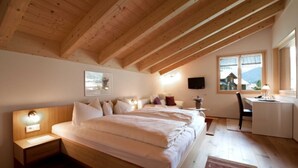 A bedroom with a wooden ceiling, a bed with white bedding, a bedside table with a lamp, a desk with a chair, and a window with a view of a mountain.