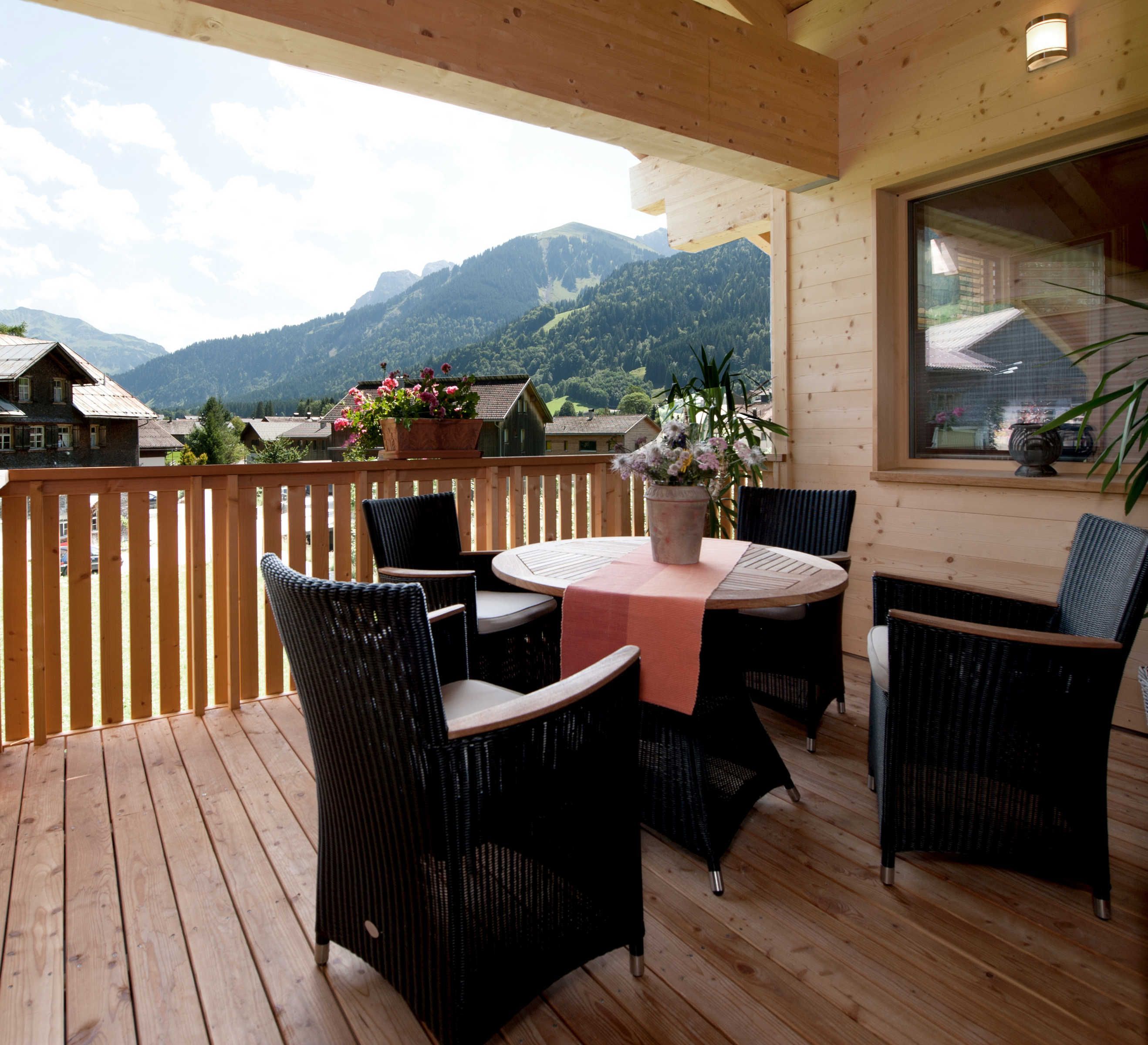 Eine Holzdiele mit Tisch und Stühlen, Blick auf Berge und ein Fenster mit Hausblick.